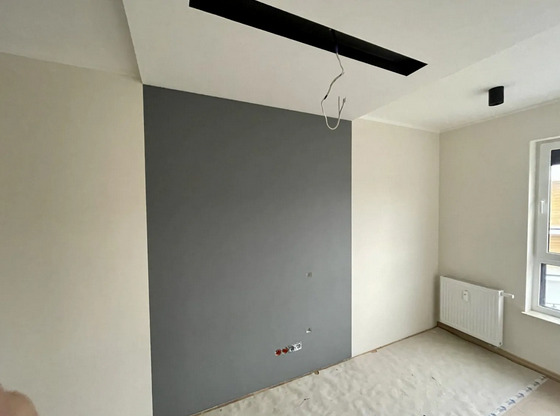 Pokój po częściowym pomalowaniu ścian: jedna ściana szara, reszta biała, widoczne gniazdka elektryczne i instalacja oświetleniowa sufitowa.