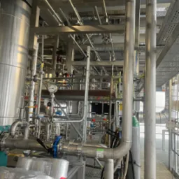 Rozbudowany system rur przemysłowych ze stali nierdzewnej z zaworami, manometrem i izolacją, widoczne elementy konstrukcji wsporczej i kraty pomostowe.