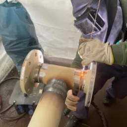 Spawanie rury z kołnierzem przez spawacza w masce ochronnej, widoczne iskry i ślady spawania, w tle worek i sprzęt spawalniczy.