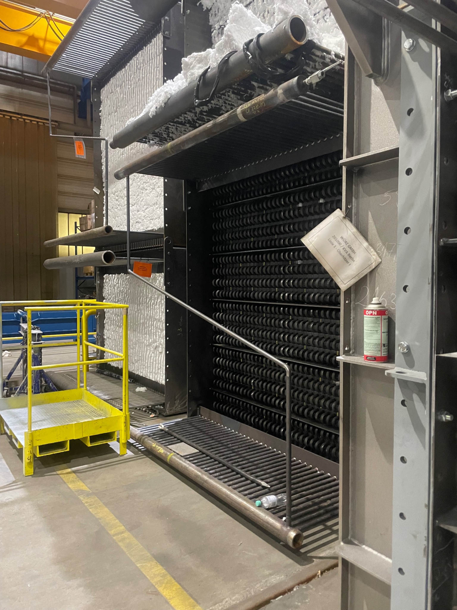 Wnętrze przemysłowego pieca z rzędami rur, izolacją z białej wełny mineralnej i konstrukcją stalową, widoczne elementy rusztowania i butelka wody na podeście.