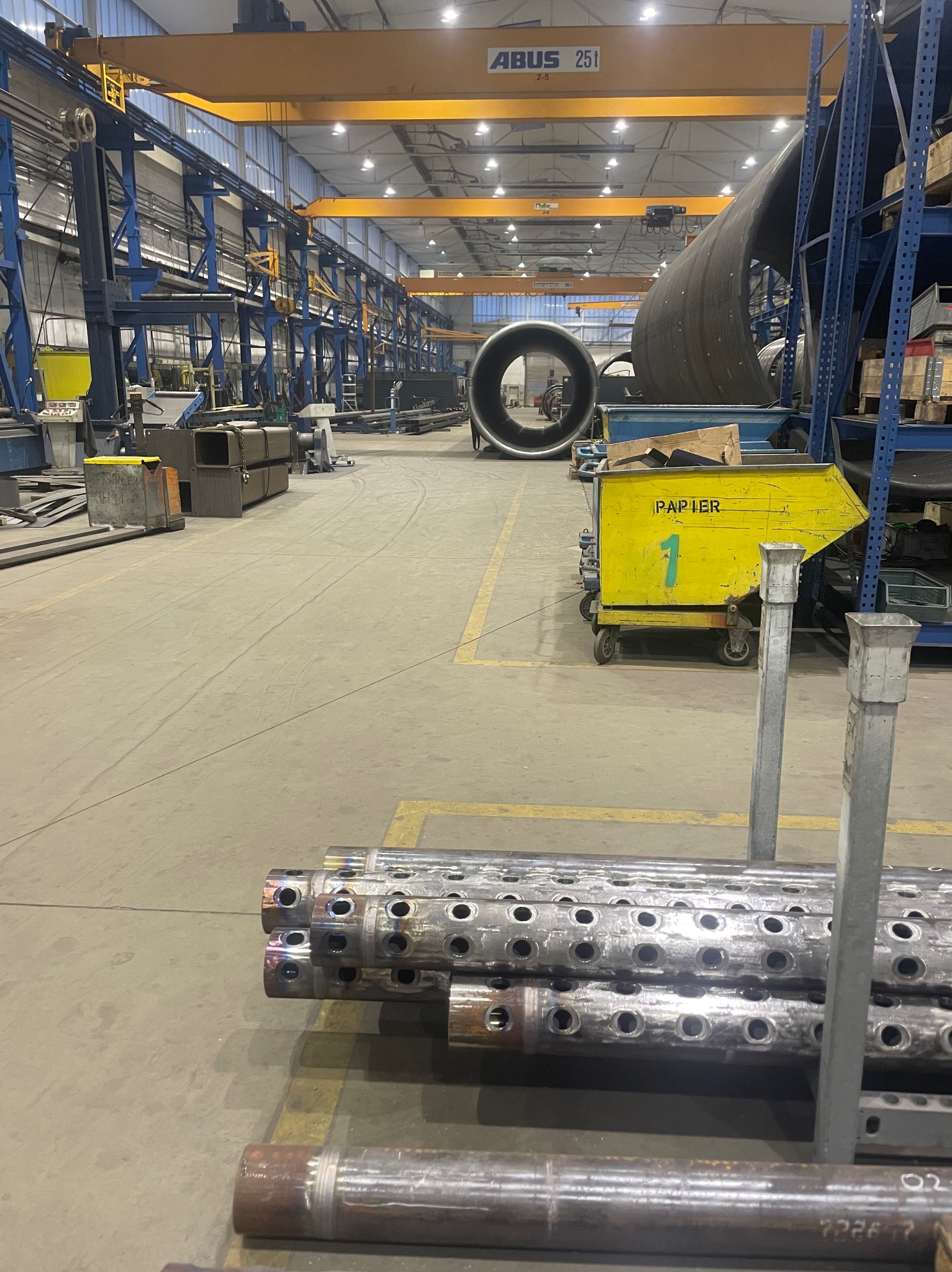 Wnętrze hali produkcyjnej z rurami metalowymi z otworami w pierwszym planie, w tle duże rury i suwnica ABUS