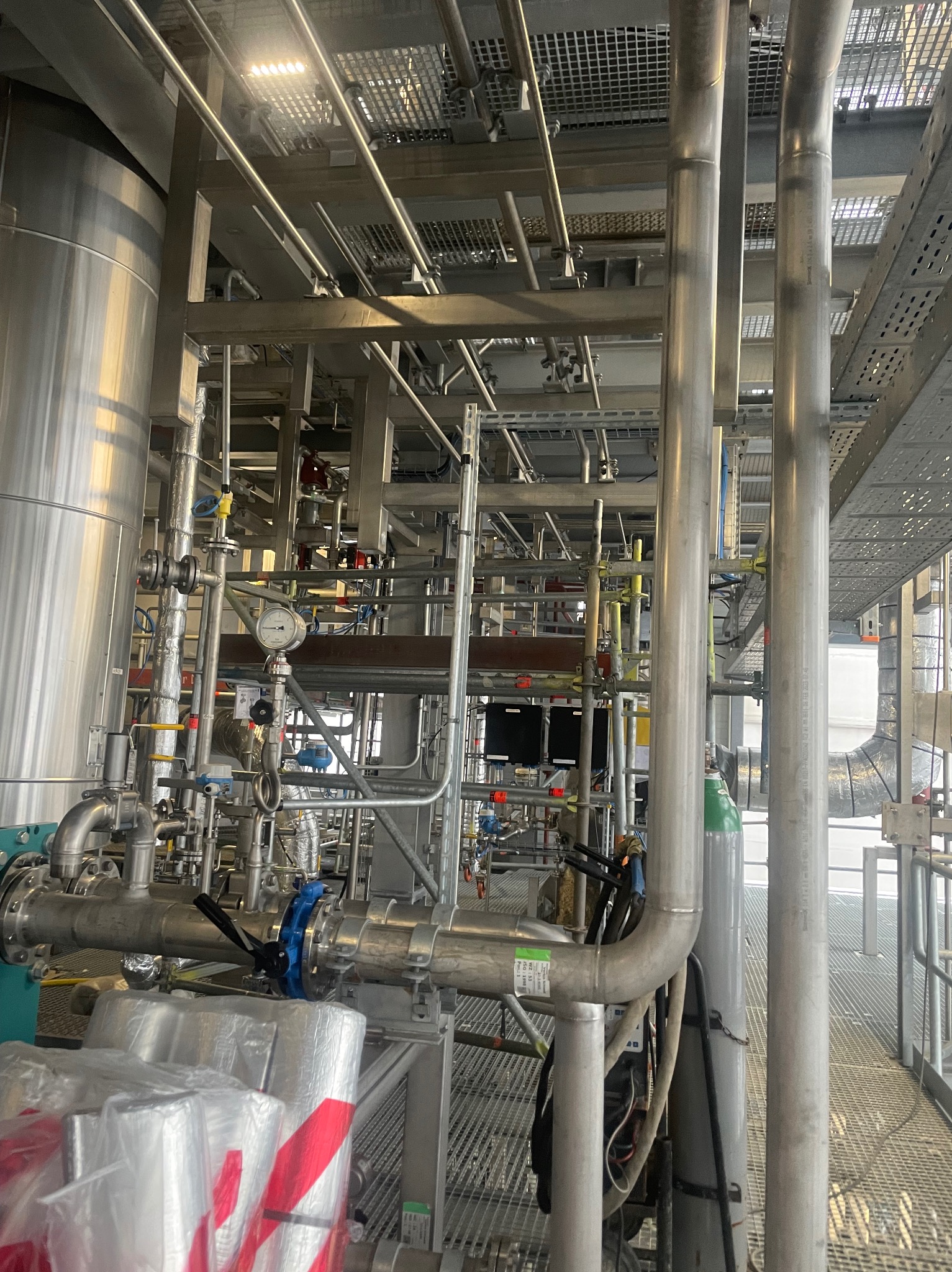 Rozbudowany system rur przemysłowych ze stali nierdzewnej z zaworami, manometrem i izolacją, widoczne elementy konstrukcji wsporczej i kraty pomostowe.