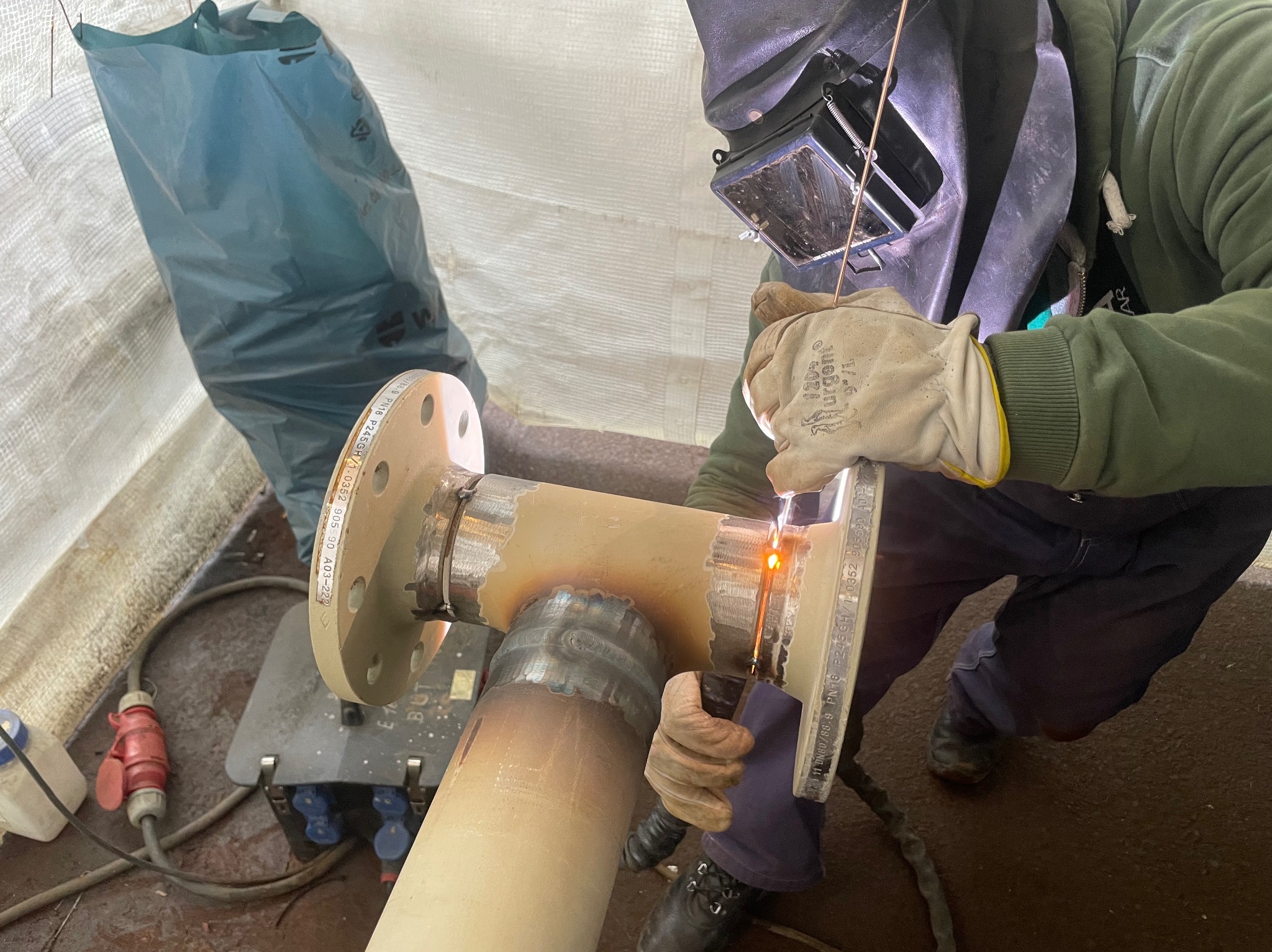 Spawanie rury z kołnierzem przez spawacza w masce ochronnej, widoczne iskry i ślady spawania, w tle worek i sprzęt spawalniczy.