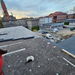 Dag-Bud S.C - Prace dekarskie na dachu budynku z widokiem na miejską zabudowę i kościół, widać palnik dekarski w użyciu oraz rolki papy z logo firmy LEMAR.