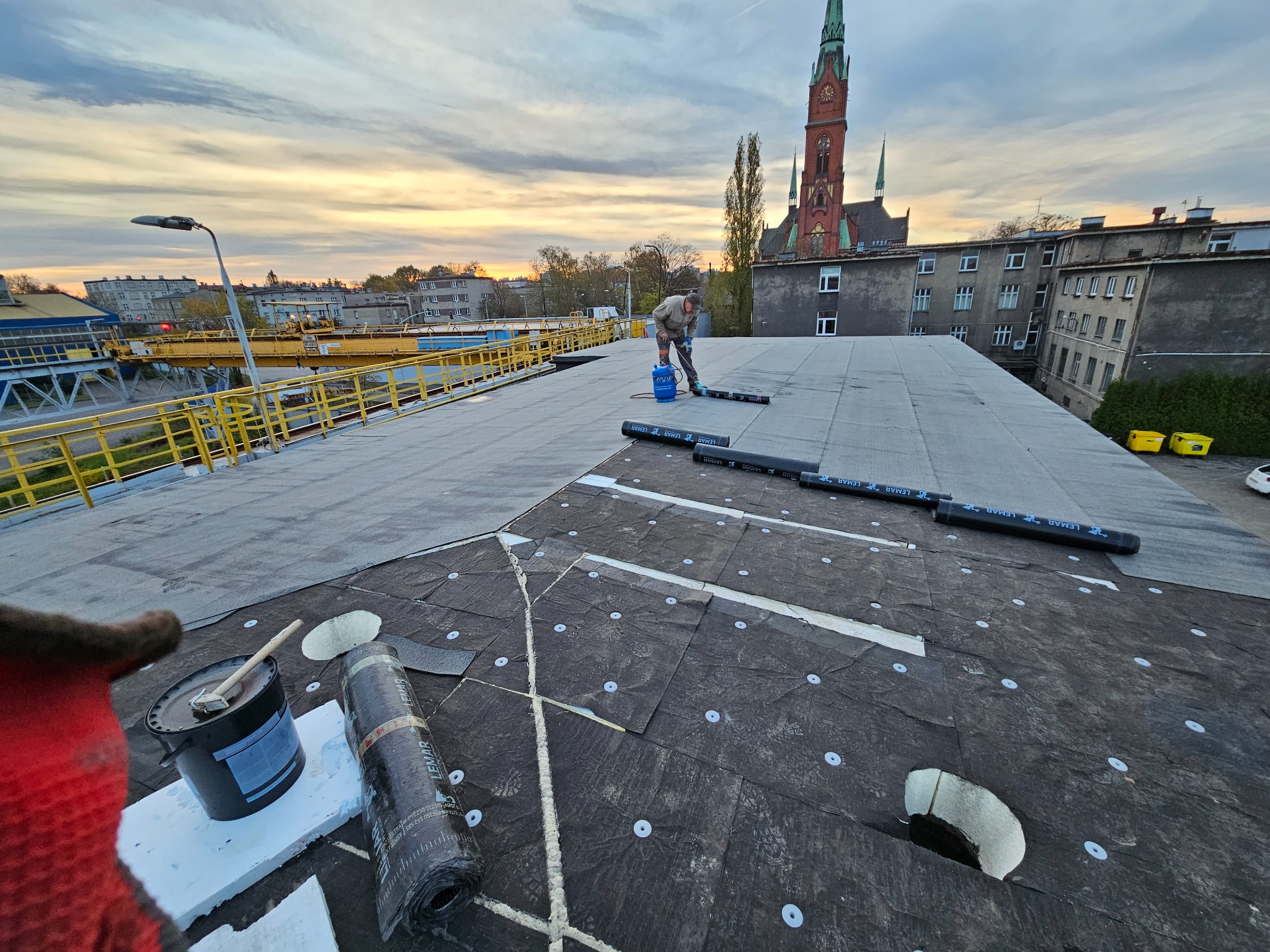 Prace na dachu budynku z widokiem na kościół w tle; robotnik używa palnika do łączenia papy termozgrzewalnej, obok leżą rolki papy marki LEMAR i wiadro z klejem.