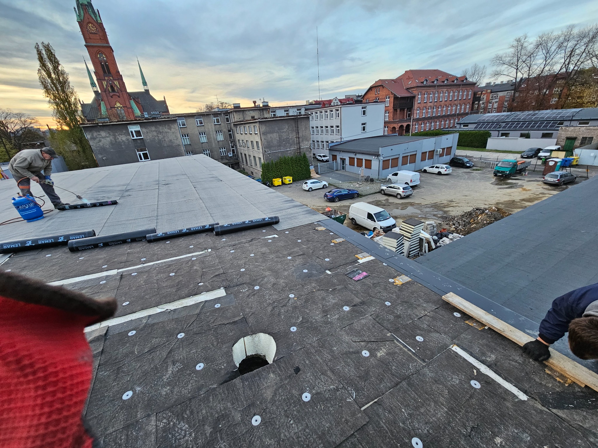 Prace dekarskie na dachu budynku z widokiem na miejską zabudowę i kościół, widać palnik dekarski w użyciu oraz rolki papy z logo firmy LEMAR.