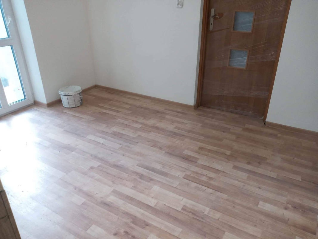 Jasne panele podłogowe ułożone w pokoju, widoczna ściana z oknem, wiadro po farbie i drewniane drzwi zabezpieczone folią.