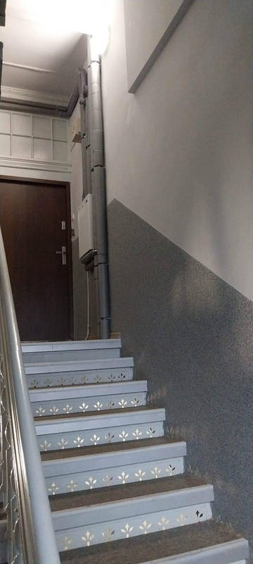 Klatka schodowa w kamienicy z szarymi ścianami i schodami z dekoracyjnymi motywami roślinnymi, pionowa rura instalacyjna biegnąca wzdłuż ściany.