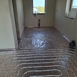 KAN-BUD Usługi wodno-kanalizacyjne i ogolnobudowlane - Białe rury ogrzewania podłogowego ułożone na folii z czerwoną siatką w surowym wnętrzu. Widoczne okno i fragment ściany z instalacją. Etap przed wylaniem posadzki.