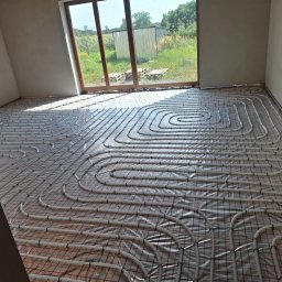 KAN-BUD Usługi wodno-kanalizacyjne i ogolnobudowlane - Białe rury ogrzewania podłogowego ułożone na folii z czerwoną siatką w surowym wnętrzu z dużym oknem i widokiem na zieleń. Etap instalacji przed wylewką.