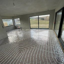 KAN-BUD Usługi wodno-kanalizacyjne i ogolnobudowlane - Nowoczesny pokój z ułożonym systemem ogrzewania podłogowego na folii izolacyjnej, widok na zewnątrz przez duże okna. Etap przed wylaniem posadzki.