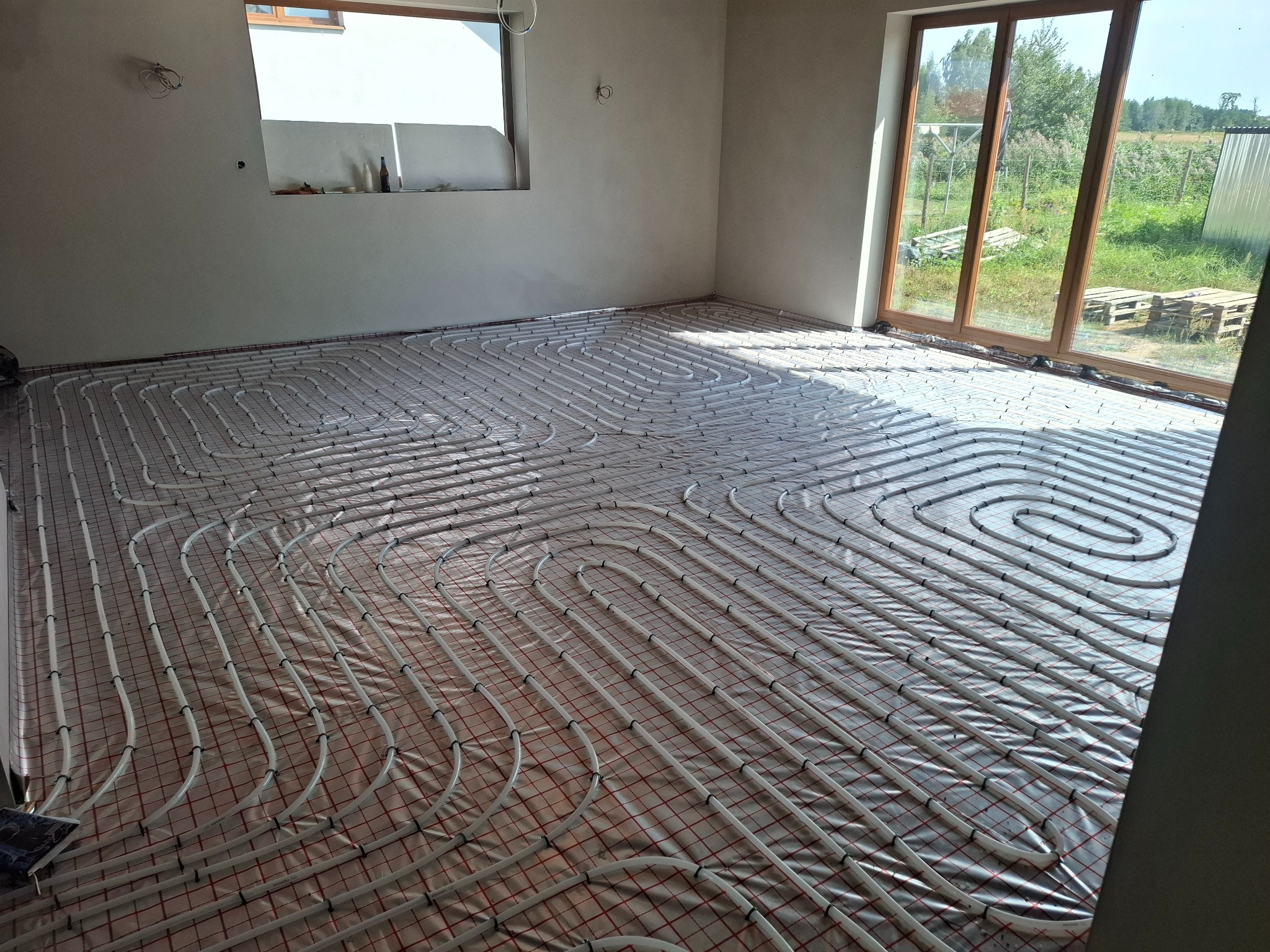 Białe rury ogrzewania podłogowego ułożone spiralnie na folii w jasnym pomieszczeniu z oknem i widokiem na zewnątrz. Widoczne mocowania rur do podłoża.