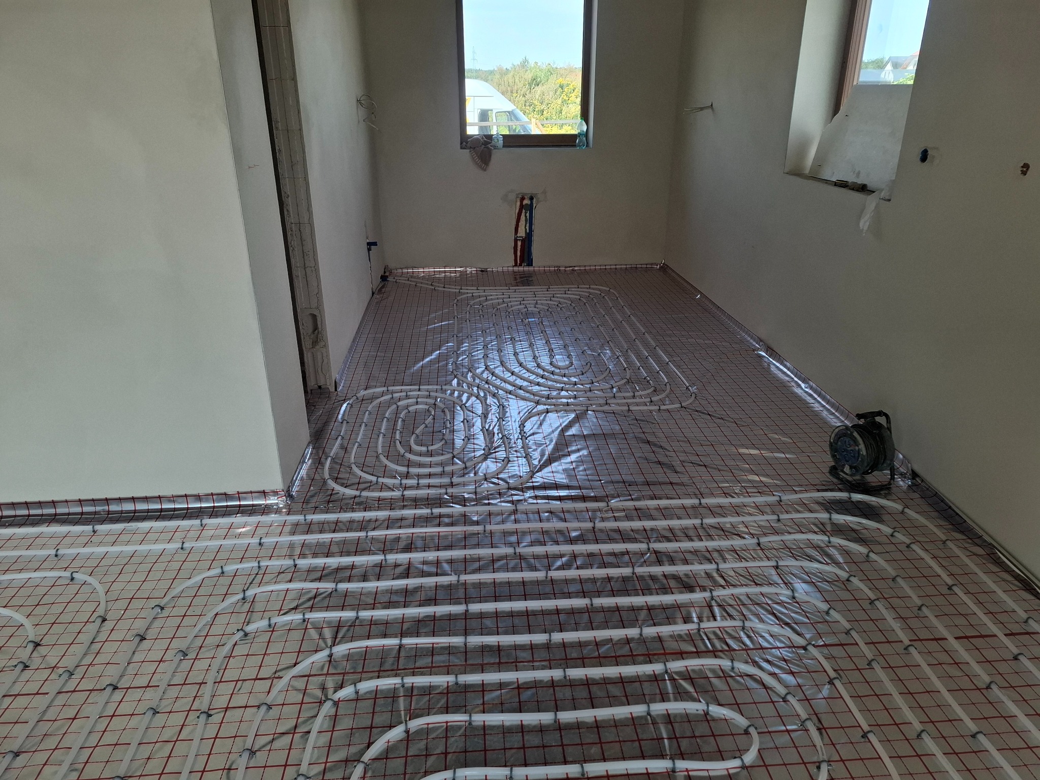 Białe rury ogrzewania podłogowego ułożone na folii z czerwoną siatką w surowym wnętrzu. Widoczne okno i fragment ściany z instalacją. Etap przed wylaniem posadzki.