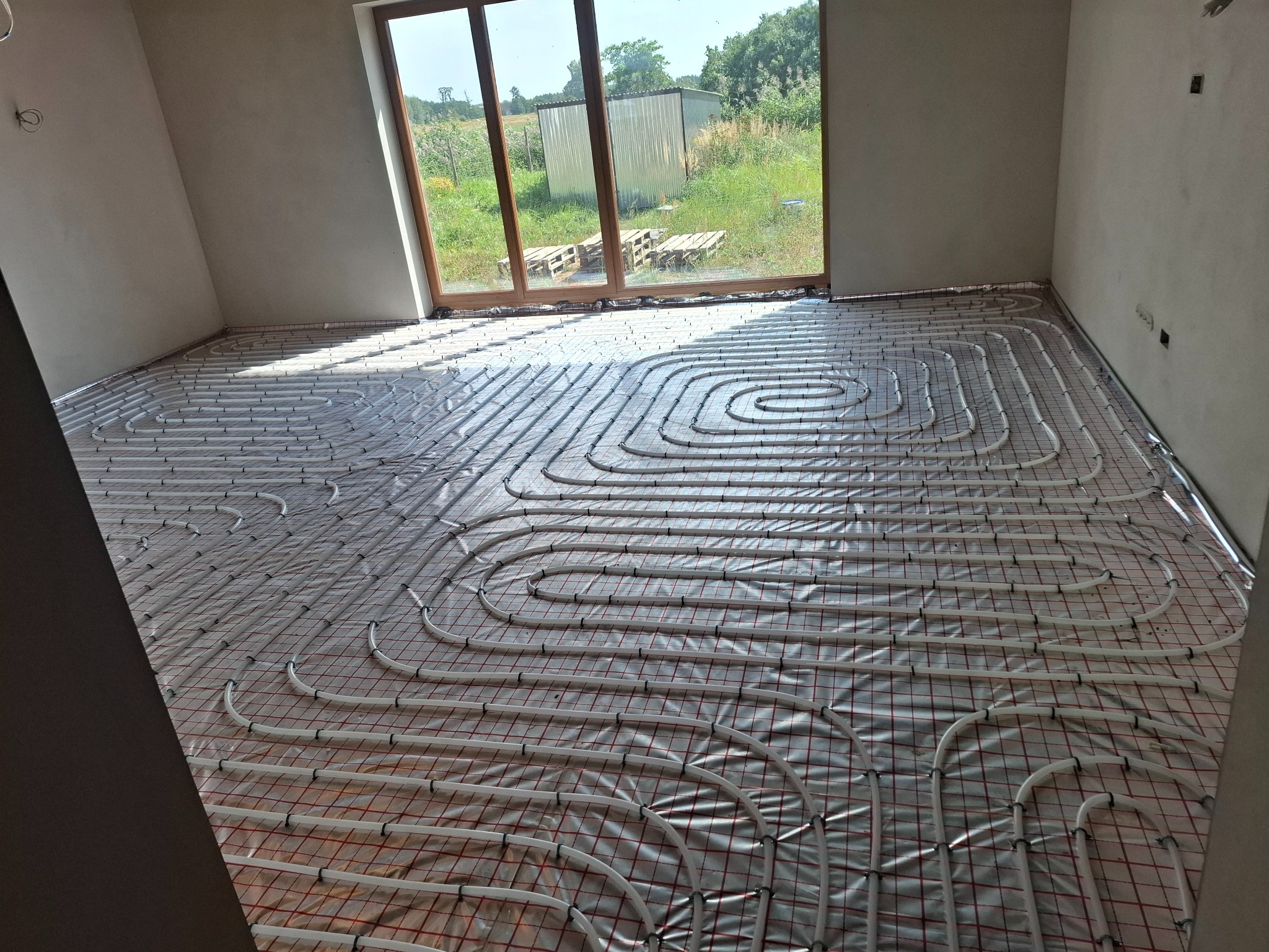 Białe rury ogrzewania podłogowego ułożone na folii z czerwoną siatką w surowym wnętrzu z dużym oknem i widokiem na zieleń. Etap instalacji przed wylewką.