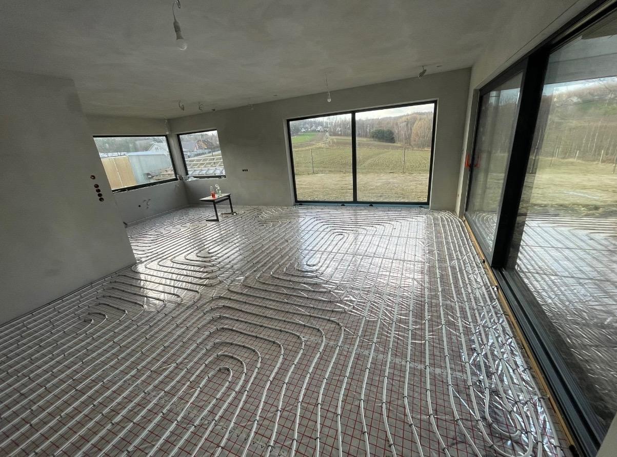 Nowoczesny pokój z ułożonym systemem ogrzewania podłogowego na folii izolacyjnej, widok na zewnątrz przez duże okna. Etap przed wylaniem posadzki.