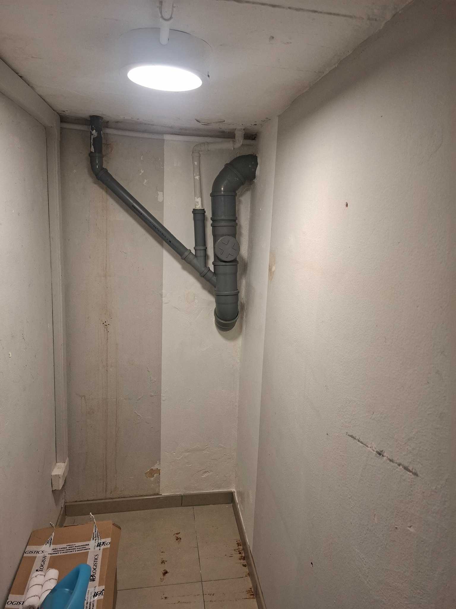 Szare rury kanalizacyjne zamontowane w rogu pomieszczenia z widocznymi śladami wilgoci na ścianach i kartonem z akcesoriami malarskimi na podłodze.