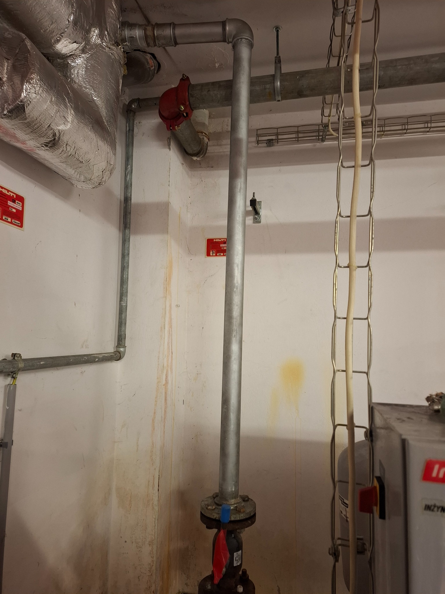 Szara pionowa rura z zaworem, fragment instalacji w pomieszczeniu technicznym. Widoczne elementy mocowań, rury poziome, ślady wilgoci na ścianie i fragment urządzenia.