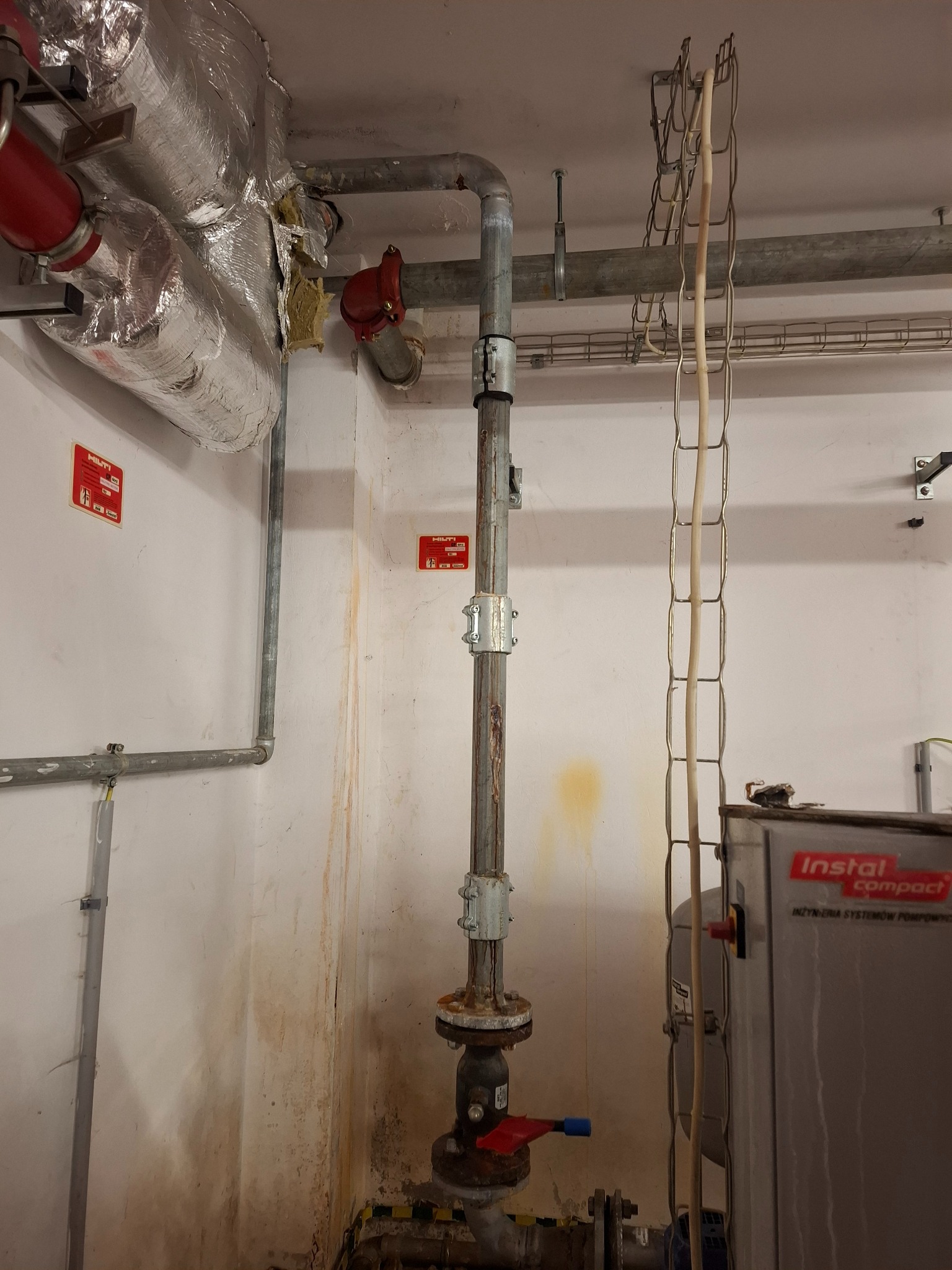 Szara pionowa rura z zaworem, zabezpieczona obejmami, biegnie od podłogi do sufitu w pomieszczeniu technicznym. Widoczne inne rury, kable i urządzenie Instal Compact.