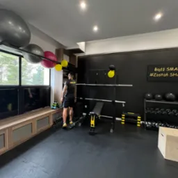 Wnętrze nowoczesnej siłowni domowej z czarną ścianą z napisem motywacyjnym 'Bądź SMART #Zostań SHAPE', wyposażonej w sztangę, ławeczkę, hantle, kettlebells, piłki gimnastyczne w różnych rozmiarach...