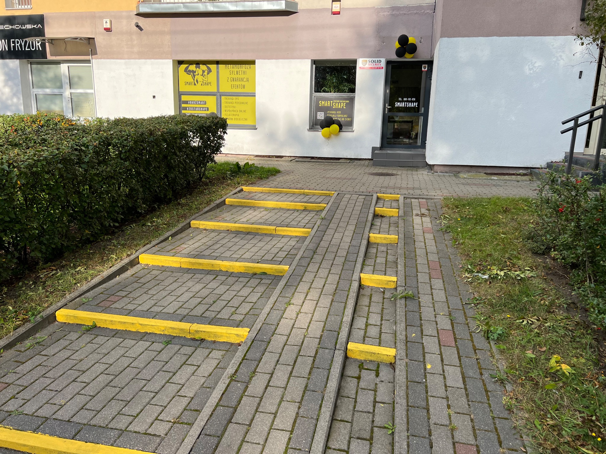 Wejście do studia treningowego Smart Shape z żółto-czarnymi balonami, rampą dla wózków i reklamami metamorfozy sylwetki na oknach.