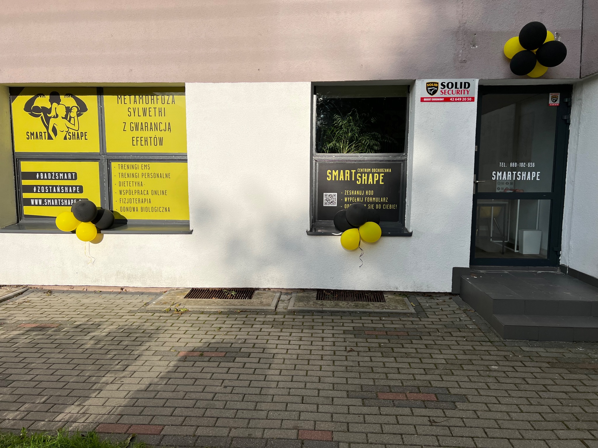 Fasada budynku centrum odchudzania SmartShape z oknami oklejonymi reklamami usług: treningi EMS, personalne, dietetyka, fizjoterapia, z czarnymi i żółtymi balonami przy oknach i drzwiach wejściowych.