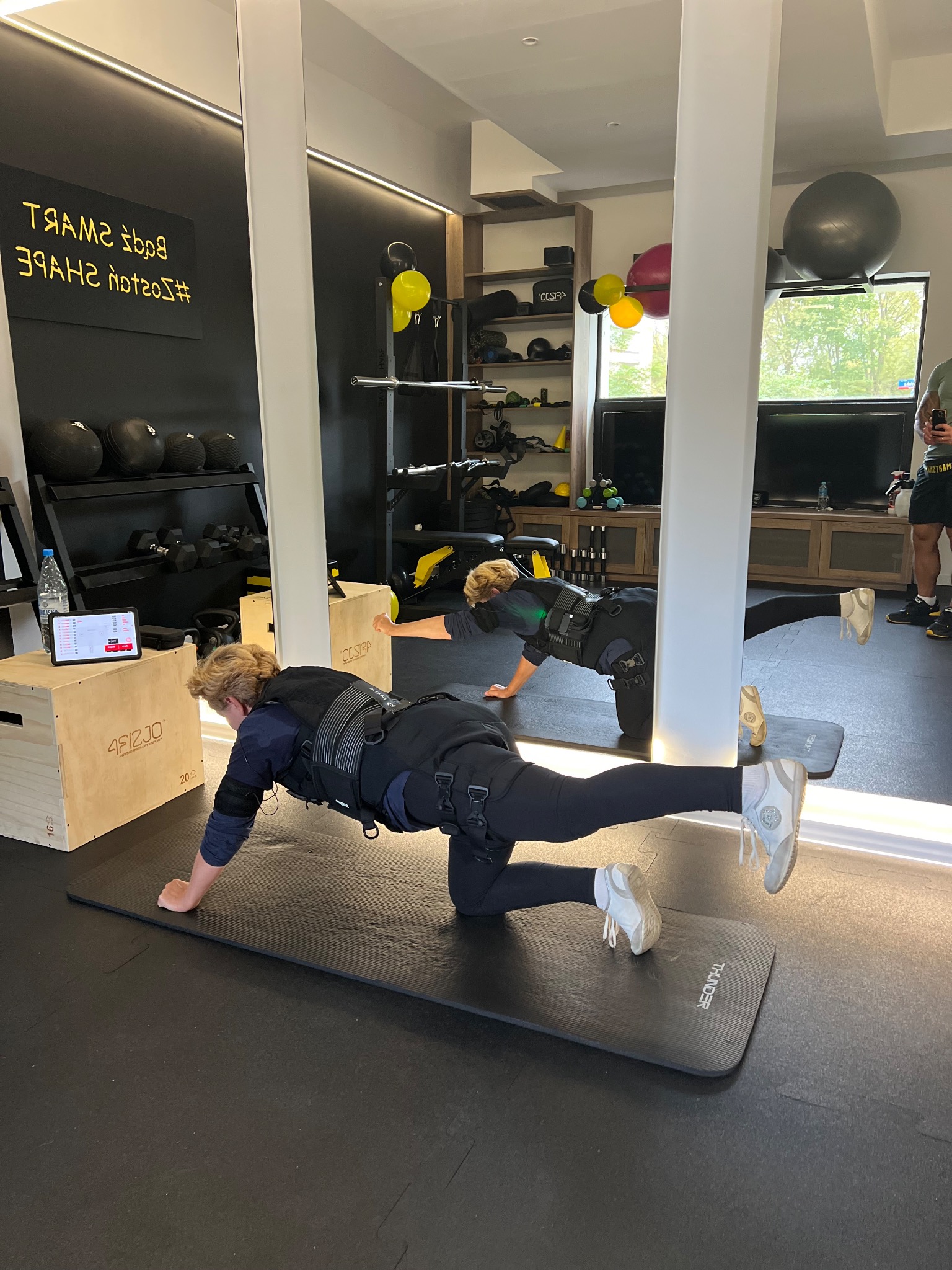 Osoba w kombinezonie EMS ćwiczy w studio, unosząc nogę do tyłu na macie; w tle lustro, sprzęt do ćwiczeń i tablica z napisem.