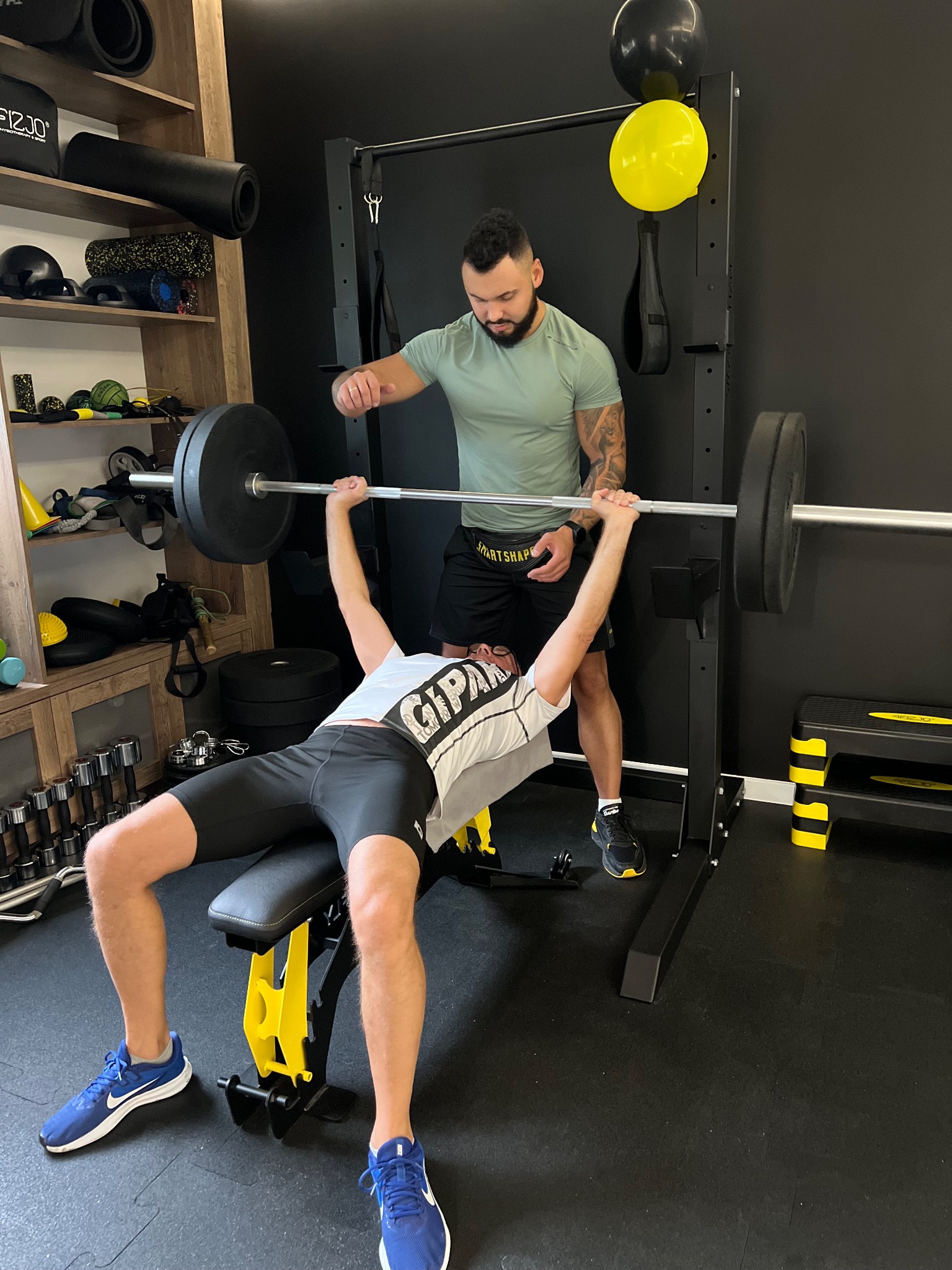 Mężczyzna ćwiczy wyciskanie sztangi leżąc na ławeczce pod nadzorem trenera w siłowni. W tle widoczne półki z akcesoriami do ćwiczeń oraz czarne i żółte balony.