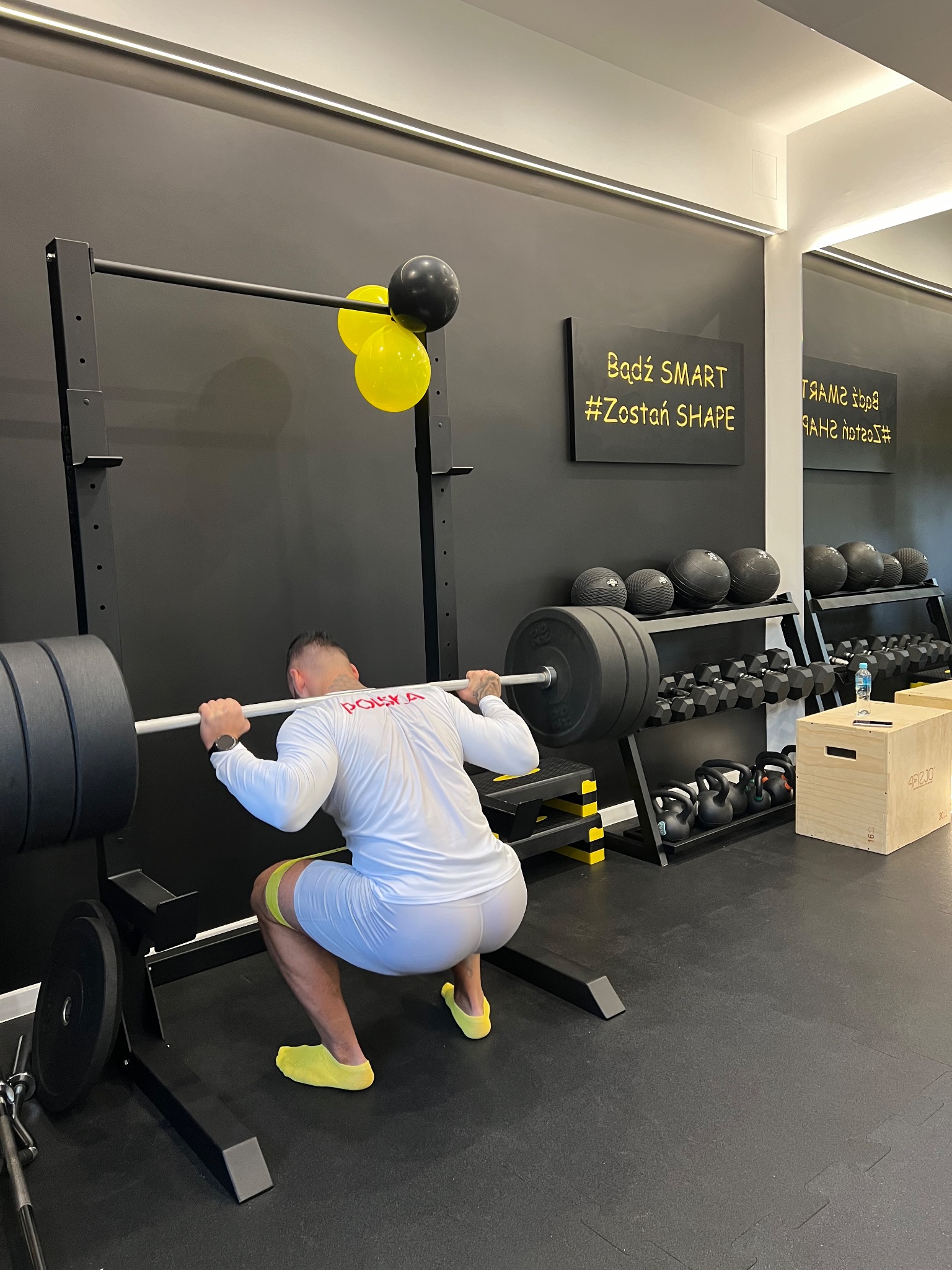 Mężczyzna w białej koszulce z napisem Polska wykonuje przysiad ze sztangą w profesjonalnej siłowni, z żółtą gumą na udach i w żółtych skarpetkach, na tle czarnej ściany z napisem 'Bądź SMART...