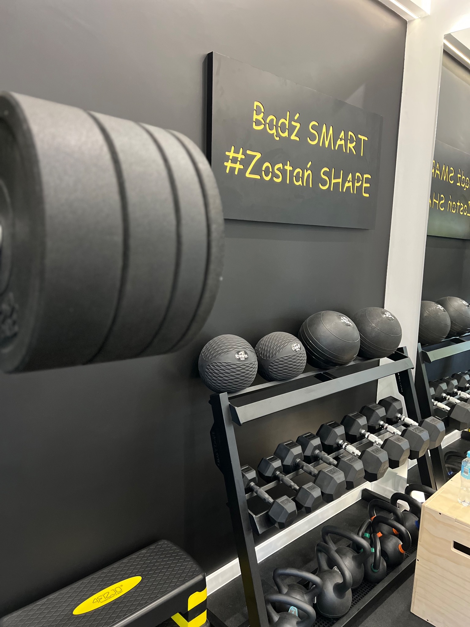 Ciężar na sztangę, piłki lekarskie, hantle i kettlebells na stojakach w siłowni z czarną ścianą i napisem 'Bądź SMART #Zostań SHAPE'.