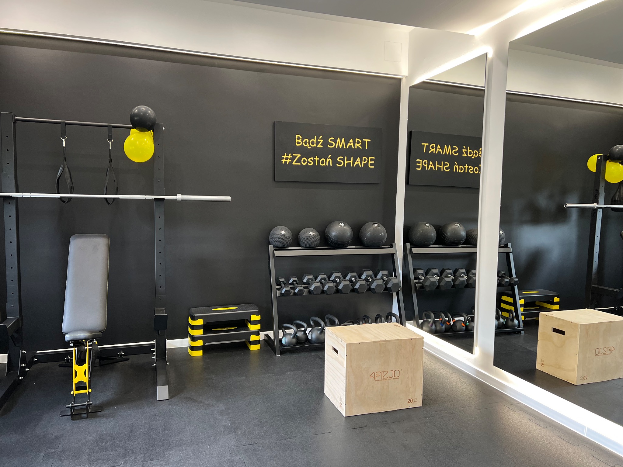 Nowoczesna siłownia z czarnymi ścianami i żółtymi akcentami, wyposażona w stojaki z hantlami, kettlebells, piłki lekarskie, ławeczkę do ćwiczeń, skrzynię do wskakiwania i napis 'Bądź SMART #Zostań...