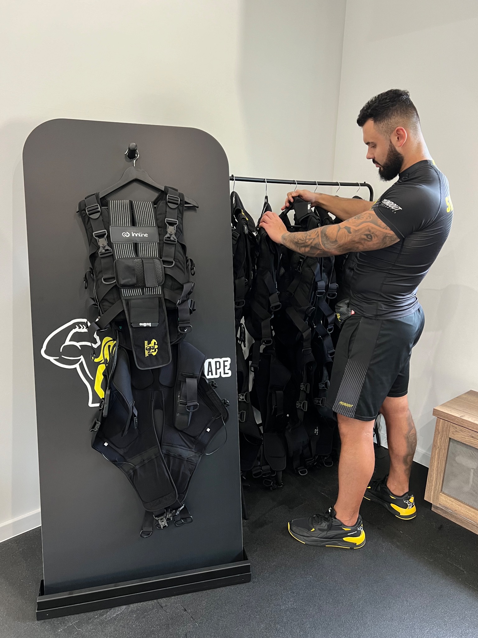 Mężczyzna z tatuażami na ramionach przygotowuje kamizelkę do elektrostymulacji mięśni, wiszącą na wieszaku w nowoczesnym studiu treningowym.
