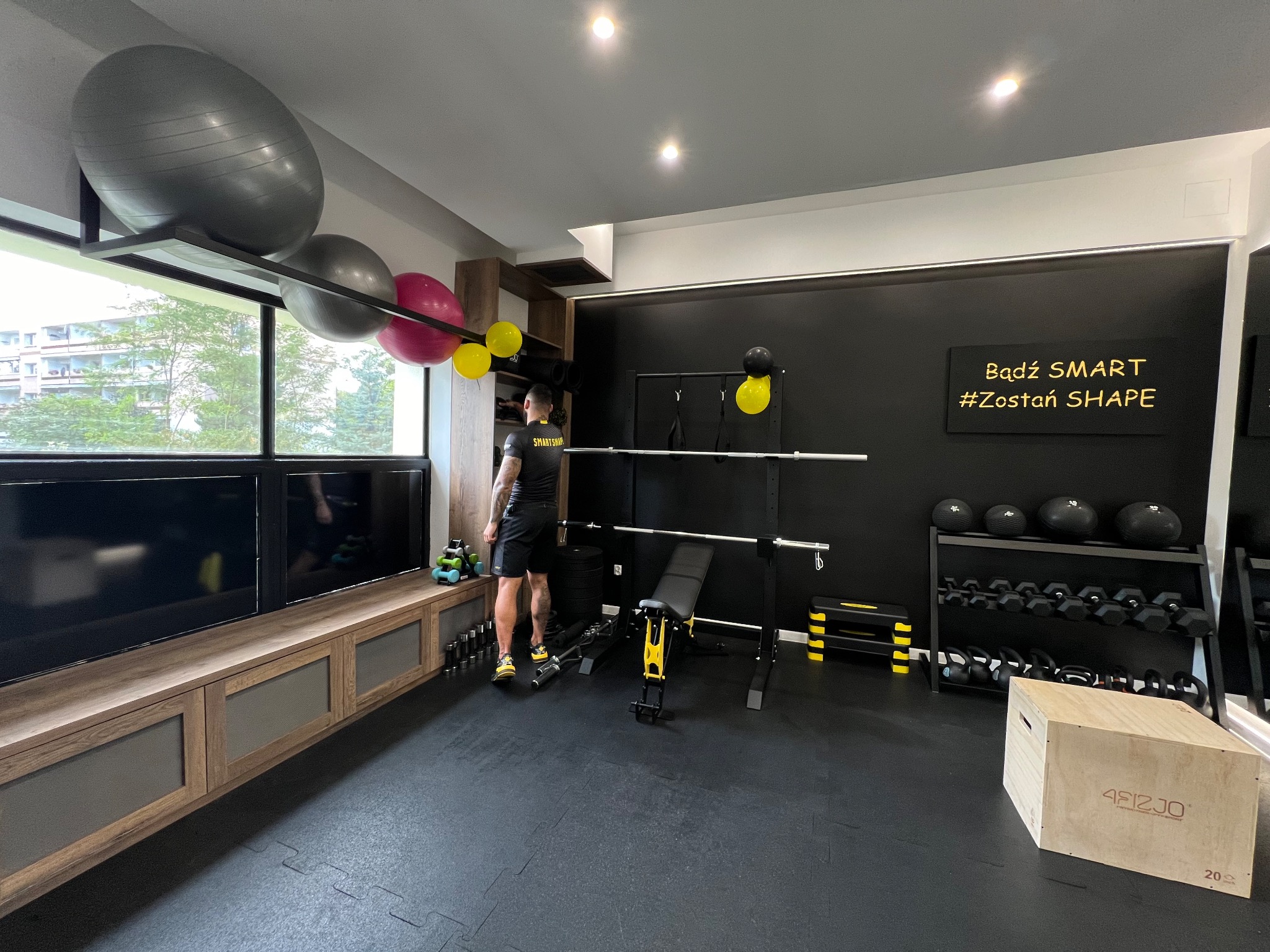 Wnętrze nowoczesnej siłowni domowej z czarną ścianą z napisem motywacyjnym 'Bądź SMART #Zostań SHAPE', wyposażonej w sztangę, ławeczkę, hantle, kettlebells, piłki gimnastyczne w różnych rozmiarach...