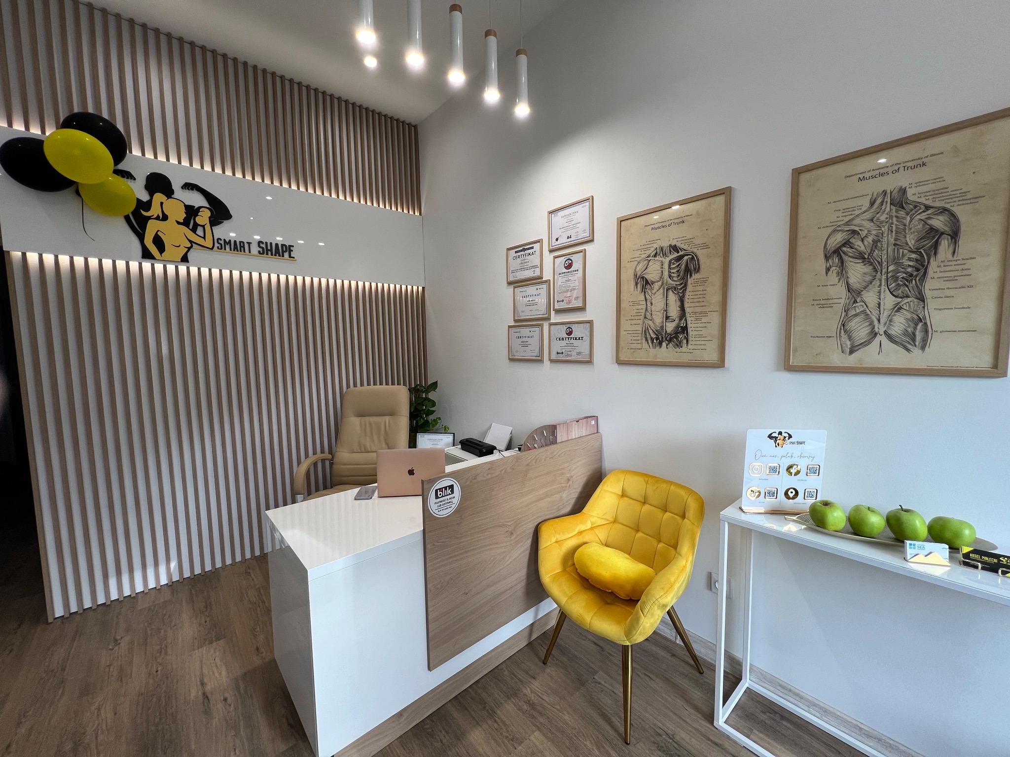 Recepcja w nowoczesnym gabinecie: biała lada, drewniane panele, żółte krzesło, dyplomy na ścianie, grafiki anatomiczne, jabłka na stole, logo Smart Shape z balonami.