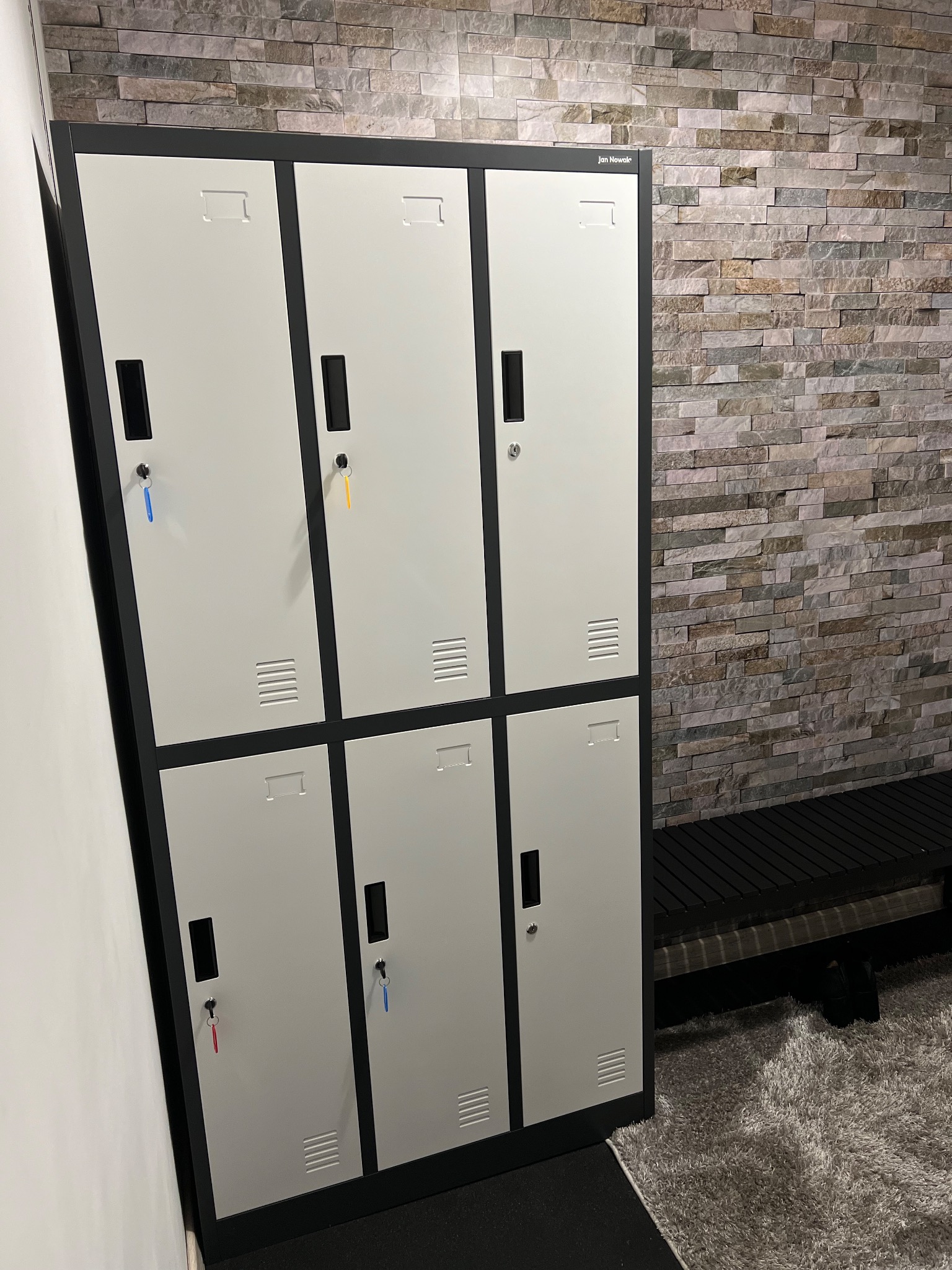Szare szafki z kluczami w siłowni w Łodzi, widoczne ławka i ściana z kamienia dekoracyjnego.