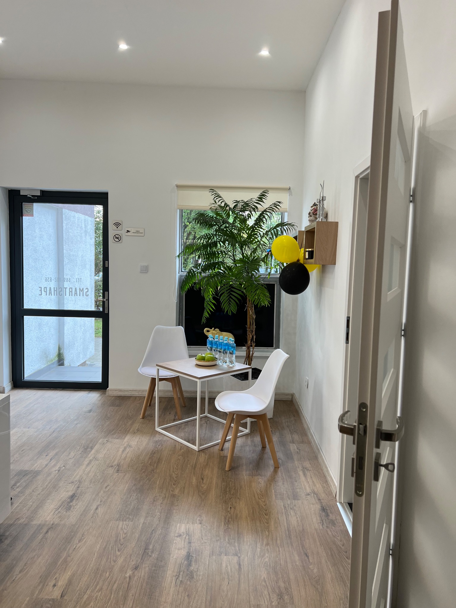 Jasne, minimalistyczne wnętrze gabinetu z białymi ścianami, drewnianą podłogą i stolikiem z krzesłami, dekorowane rośliną w doniczce, wodą mineralną i jabłkami, z czarno-żółtymi balonami na ścianie.