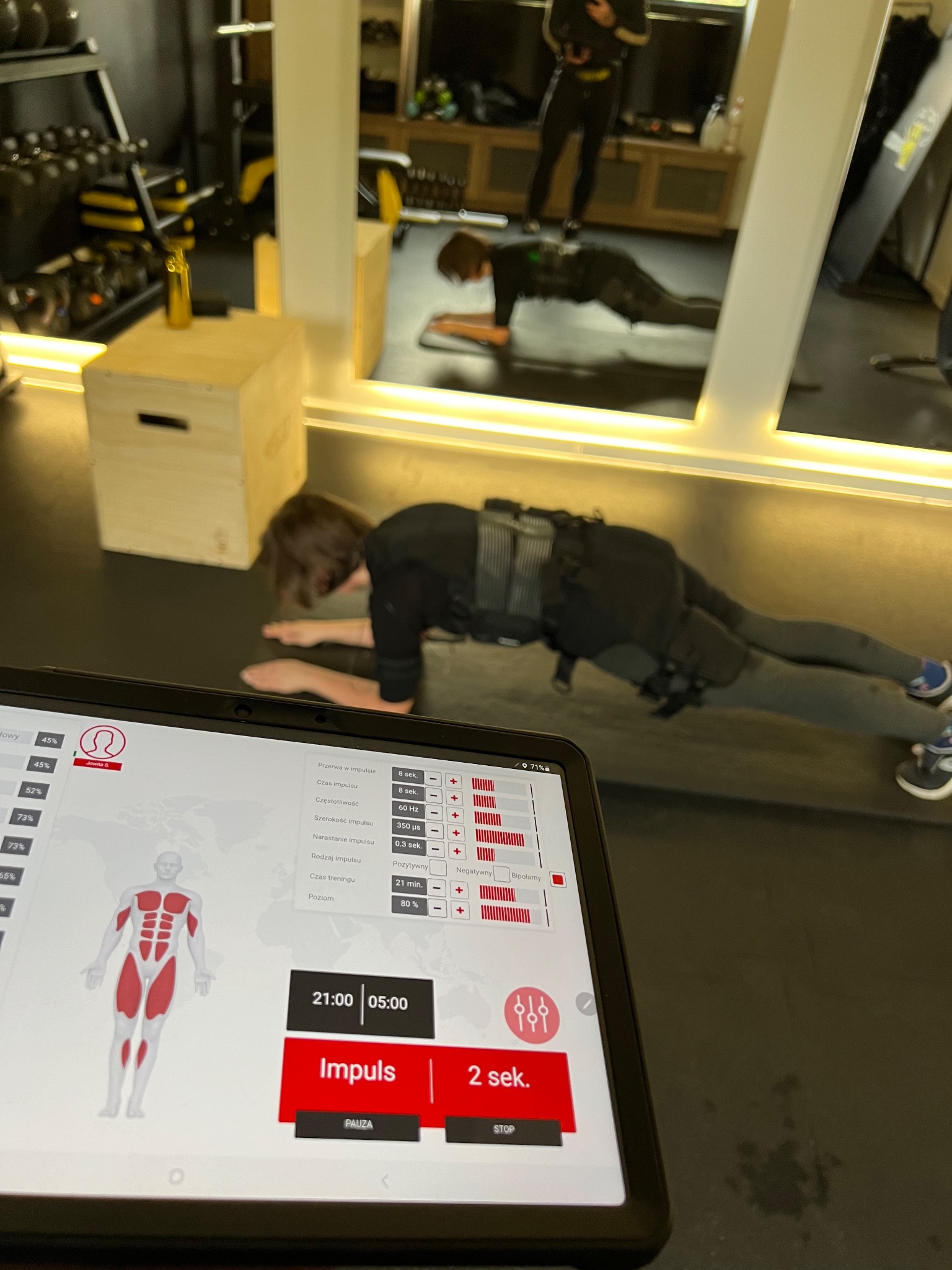 Osoba w kombinezonie EMS wykonuje ćwiczenie deski, widoczny ekran tabletu z ustawieniami impulsów elektrycznych i schematem mięśni, w tle lustro z odbiciem drugiej osoby ćwiczącej.