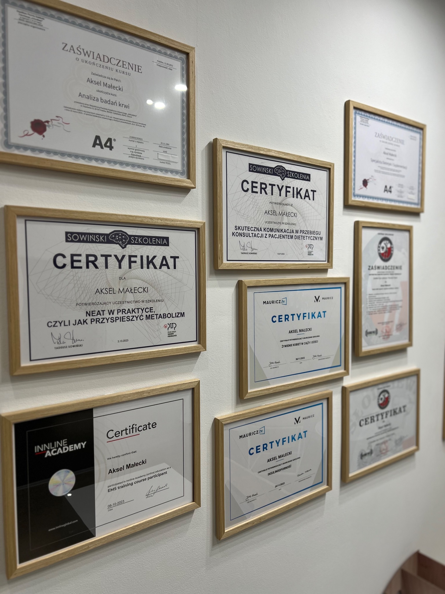 Ściana z ramkami zawierającymi certyfikaty i zaświadczenia ukończenia kursów i szkoleń, w tym certyfikaty z zakresu dietetyki, żywienia kobiet w ciąży i dzieci, analizy badań krwi oraz treningu EMS.