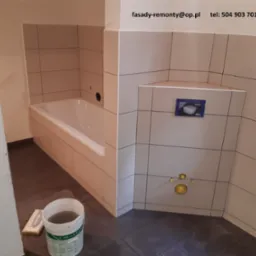 Wykończona łazienka z beżowymi płytkami, widoczna wanna, zabudowa stelaża WC Geberit, przyłącza wodne i wiadro z wodą na ciemnej podłodze.