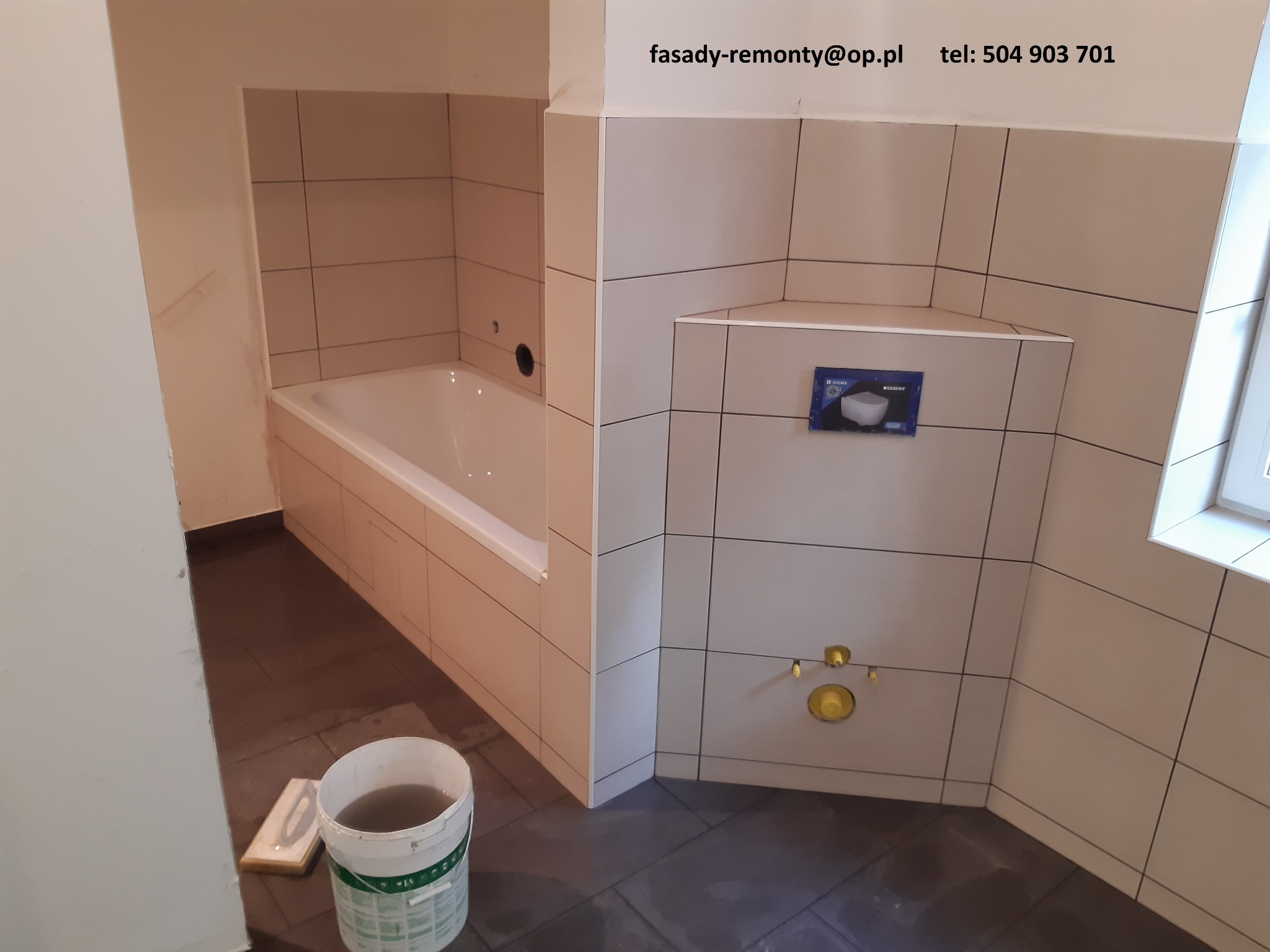 Wykończona łazienka z beżowymi płytkami, widoczna wanna, zabudowa stelaża WC Geberit, przyłącza wodne i wiadro z wodą na ciemnej podłodze.