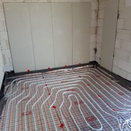 Hydroprojekt - Białe rury systemu ogrzewania podłogowego Roth ułożone na folii refleksyjnej z czerwoną siatką, widoczne czerwone klipsy mocujące, ściany z białych bloczków betonu komórkowego.