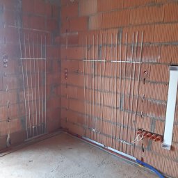 Hydroprojekt - Surowy stan wnętrza z widocznymi pionowymi rurkami instalacji grzewczej zamocowanymi do ściany z cegły dziurawki, oraz instalacją elektryczną i rurami instalacji wodnej w podłodze.