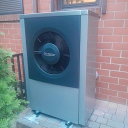 Hydroprojekt - Szara pompa ciepła Buderus zainstalowana przy ceglanym budynku, widoczna z boku, z zielonym krzewem po lewej stronie i oknem w tle.