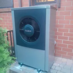 Szara pompa ciepła Buderus zainstalowana przy ceglanym budynku, widoczna z boku, z zielonym krzewem po lewej stronie i oknem w tle.