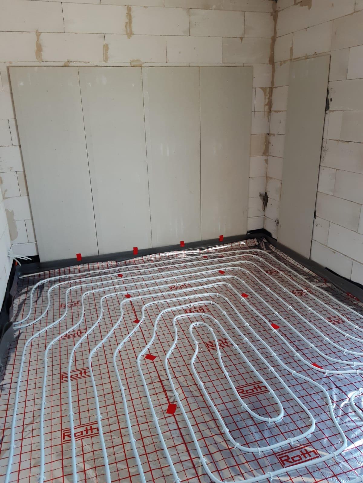 Białe rury systemu ogrzewania podłogowego Roth ułożone na folii refleksyjnej z czerwoną siatką, widoczne czerwone klipsy mocujące, ściany z białych bloczków betonu komórkowego.