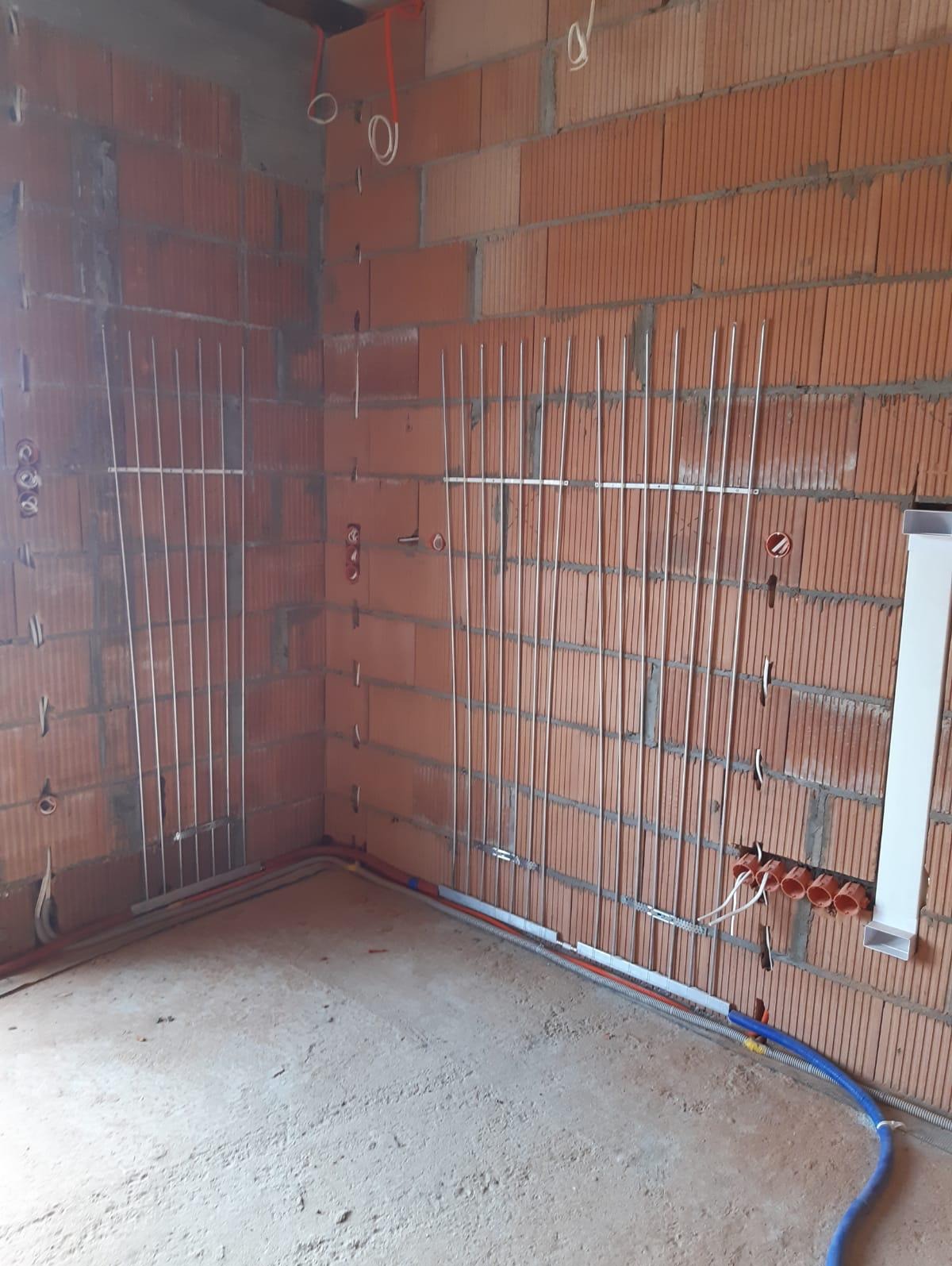 Surowy stan wnętrza z widocznymi pionowymi rurkami instalacji grzewczej zamocowanymi do ściany z cegły dziurawki, oraz instalacją elektryczną i rurami instalacji wodnej w podłodze.