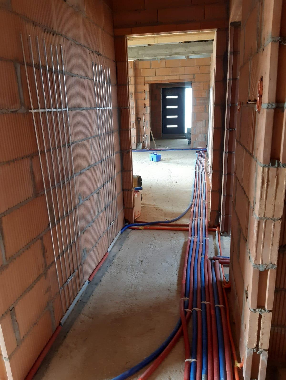 Wnętrze w trakcie budowy z widocznymi rurami instalacji wodnej (czerwone i niebieskie) ułożonymi wzdłuż podłogi oraz pionowymi rurkami na ścianie, prawdopodobnie pod ogrzewanie podłogowe.