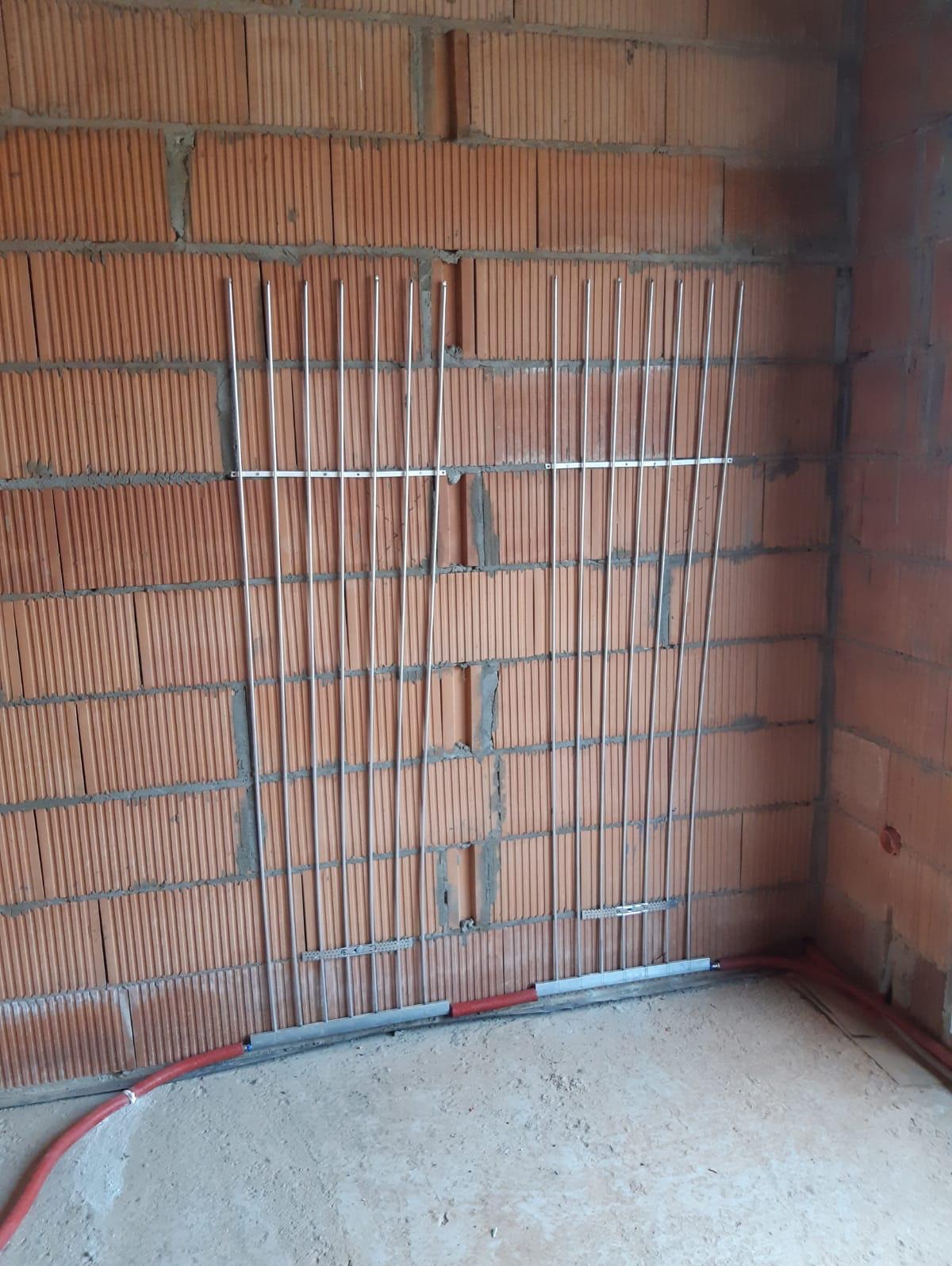 Montaż rurek instalacji ogrzewania podłogowego na ścianie z cegły dziurawki, widoczne mocowania i fragment rury w podłodze.
