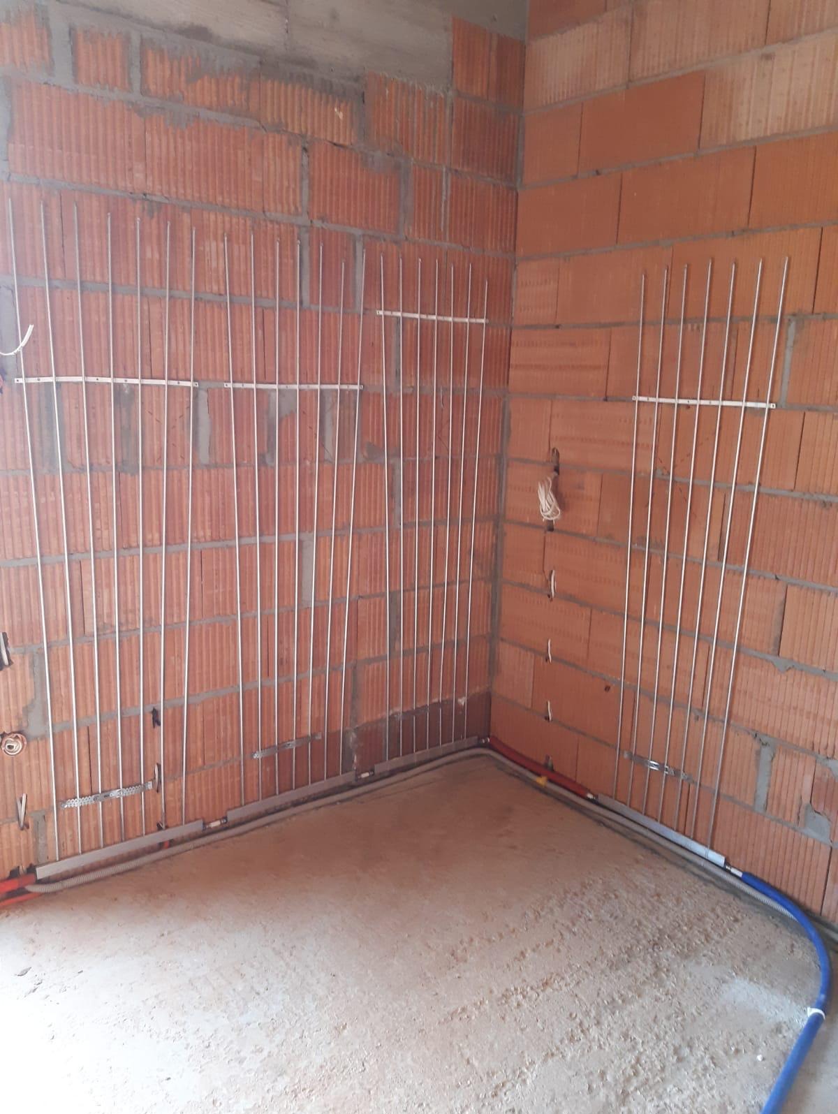Montaż ogrzewania podłogowego w surowym stanie, widoczne rury grzewcze i mocowania na ceglanych ścianach, z elementami instalacji biegnącymi wzdłuż podłogi.