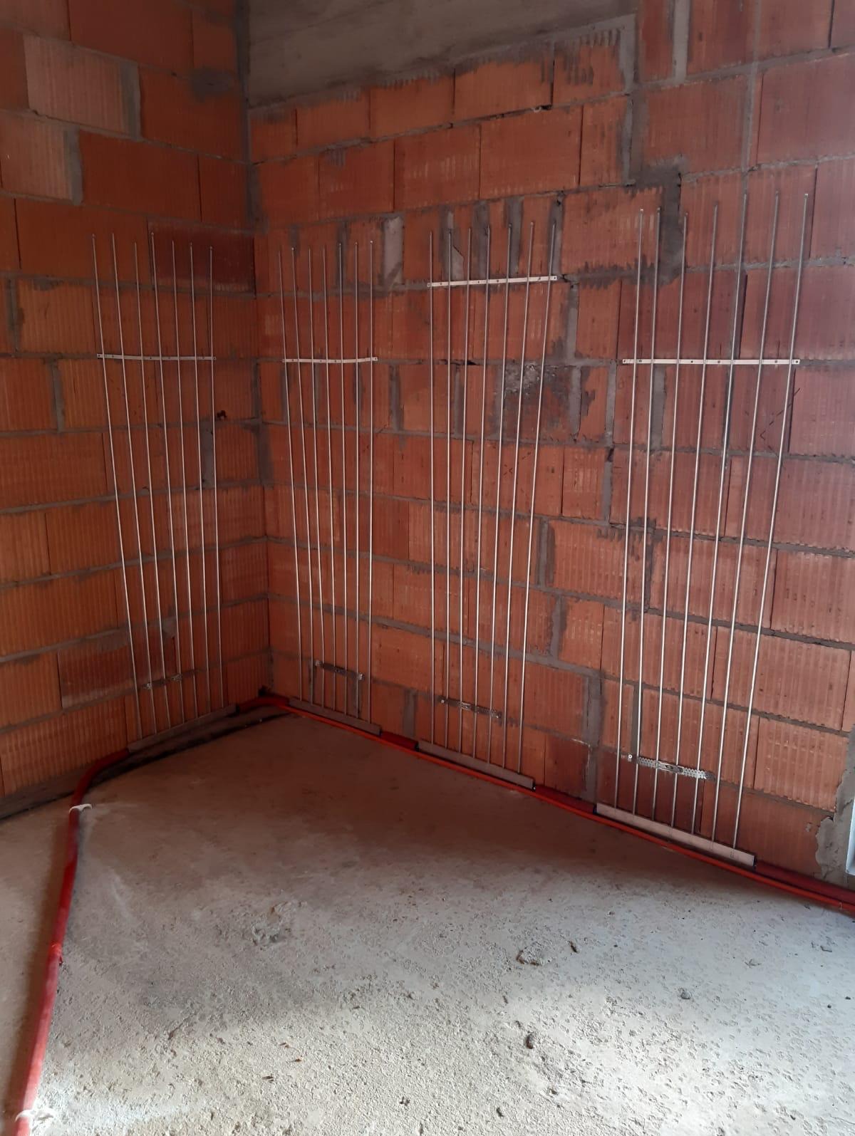 Ściana z cegły dziurawki z zamocowanymi pionowymi rurkami ogrzewania podłogowego ściennego oraz poziomymi rurkami w podłodze w surowym stanie.