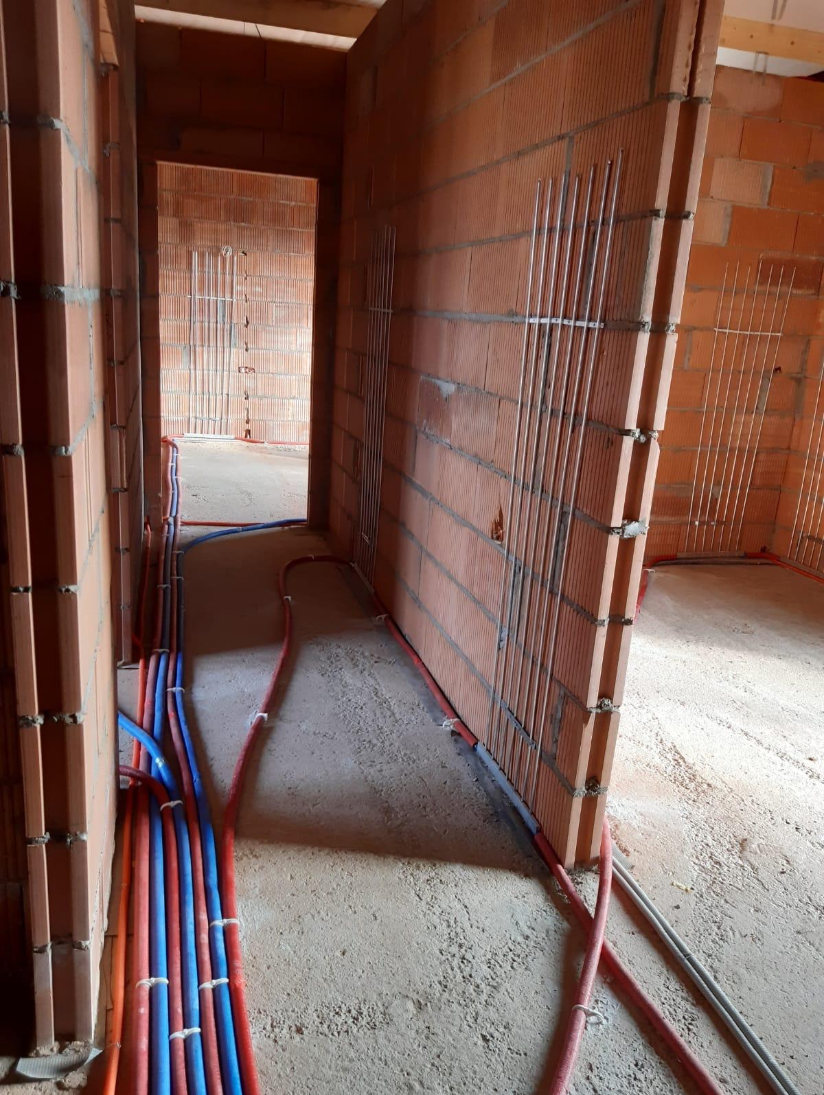 Instalacja rur grzewczych i wodnych w surowym stanie, widoczne pionowe rury w ścianach z cegły dziurawki i poziome rury na podłodze, zabezpieczone opaskami.